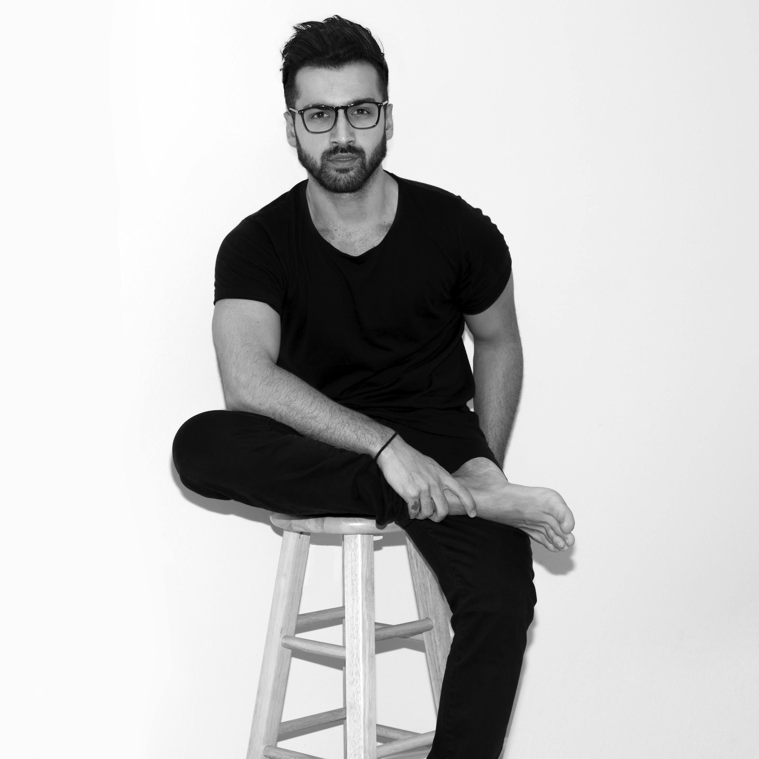 Black and white photo of a man sitting on a stool indoors, looking pensive and fashionable.